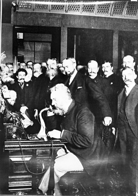 Alexander Graham Bell sits at a desk speaking on a telephone